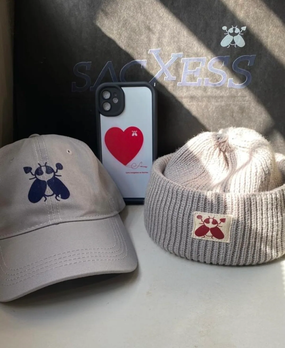 Sacxess brand-world flat lay in window light — cap, beanie and phone case